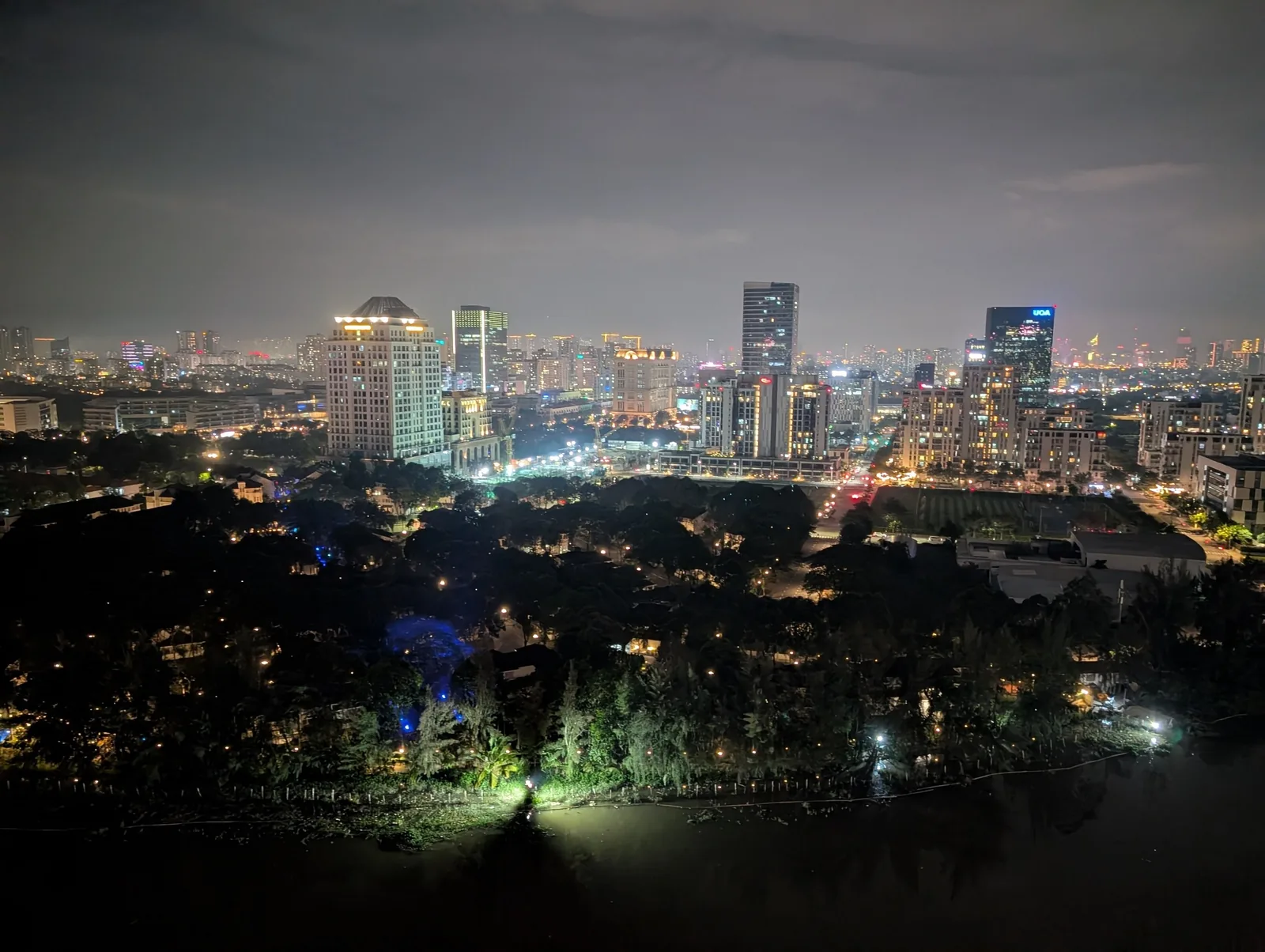 City skyline of Ho Chi Minh City glows at night above a dark tree-lined lake and quiet park.