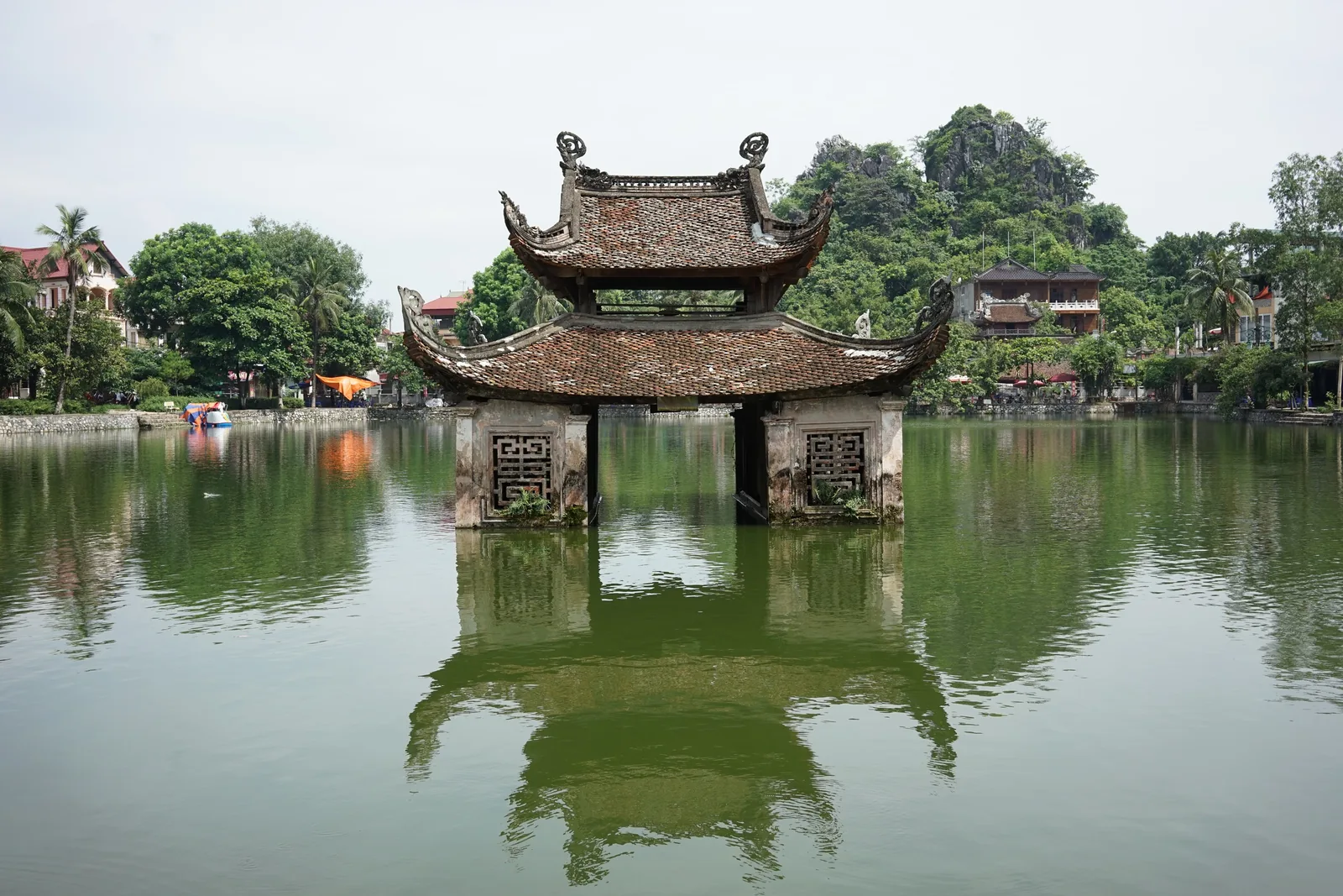 A traditional water pavilion with curved tiled roofs stands on stilts in a calm lake near Hanoi, limestone hills rising.