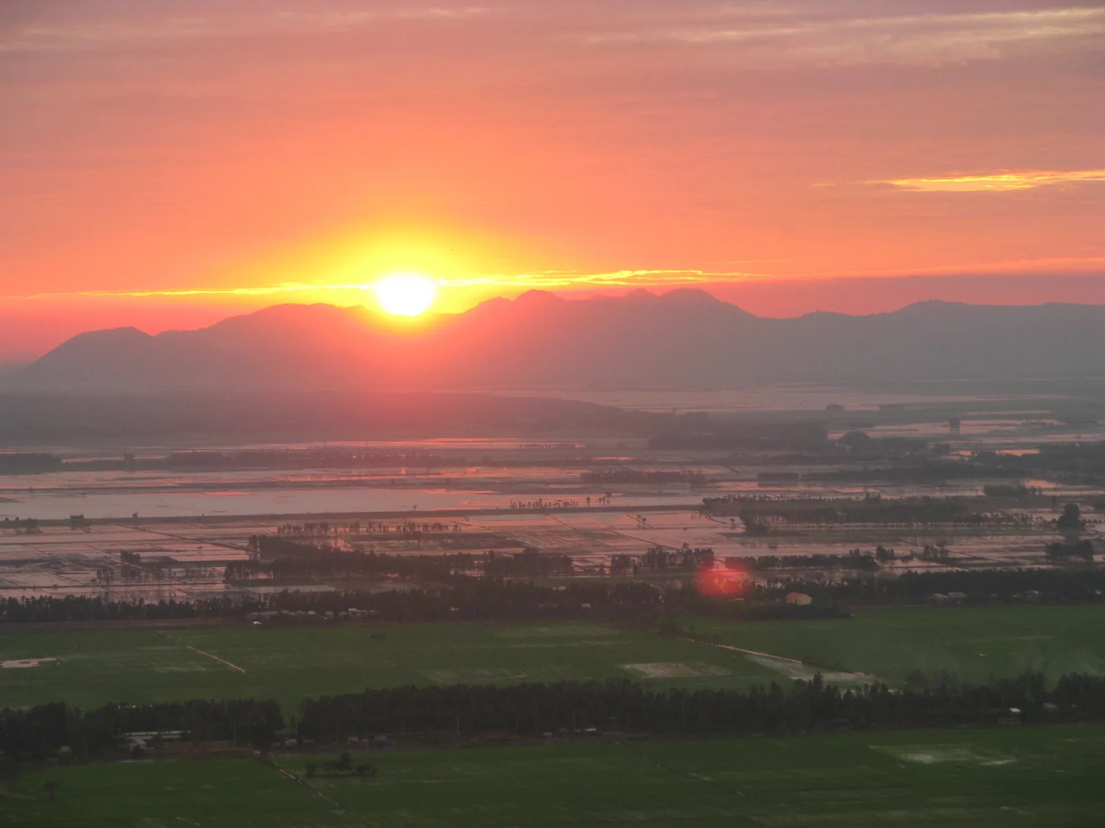 A vivid orange sunset glows behind mountains over the flooded rice fields of the Mekong Delta.