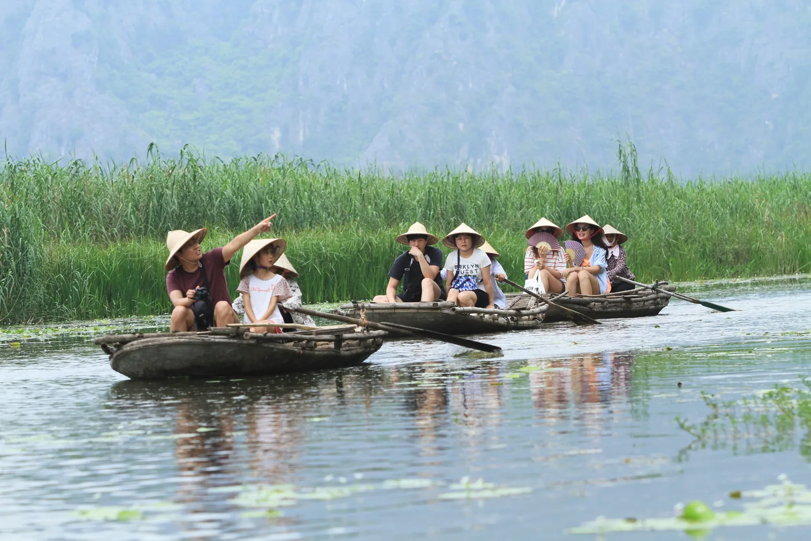 Tourists in conical hats ride small rowboats through a reed-lined river near limestone hills in Vietnam.