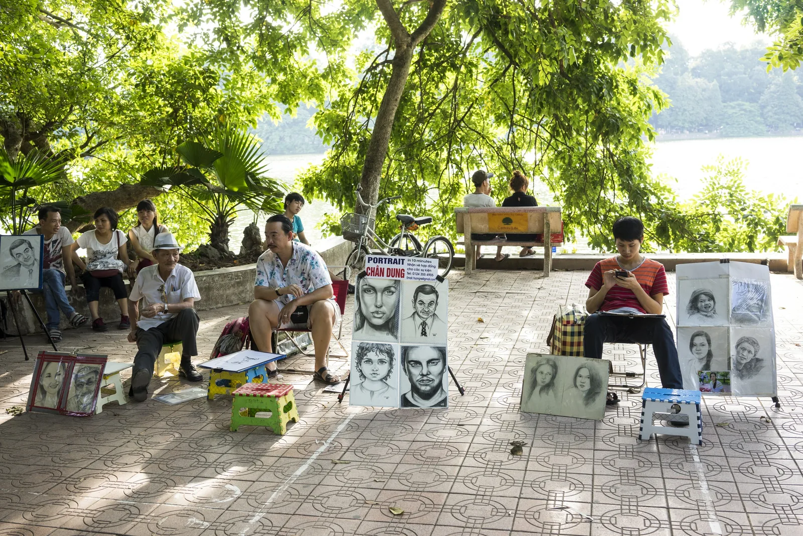 Street portrait artists display sample sketches while sitting along a shaded lakeside path in Hanoi.
