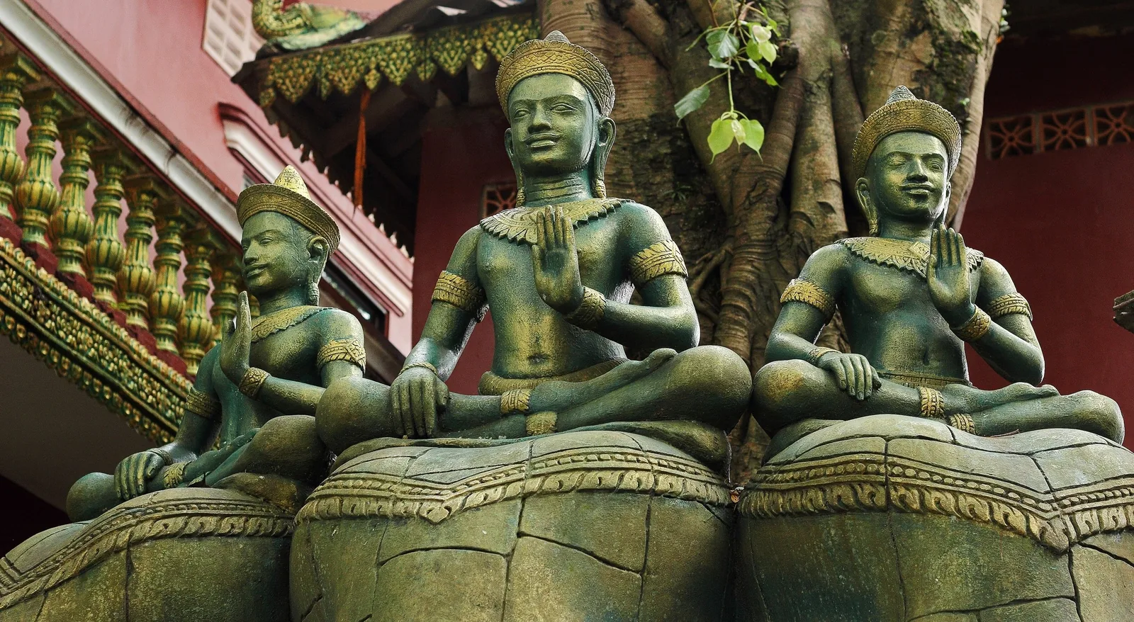 Bronze statues of seated deities with golden crowns sit atop lotus pedestals at a Cambodian temple.