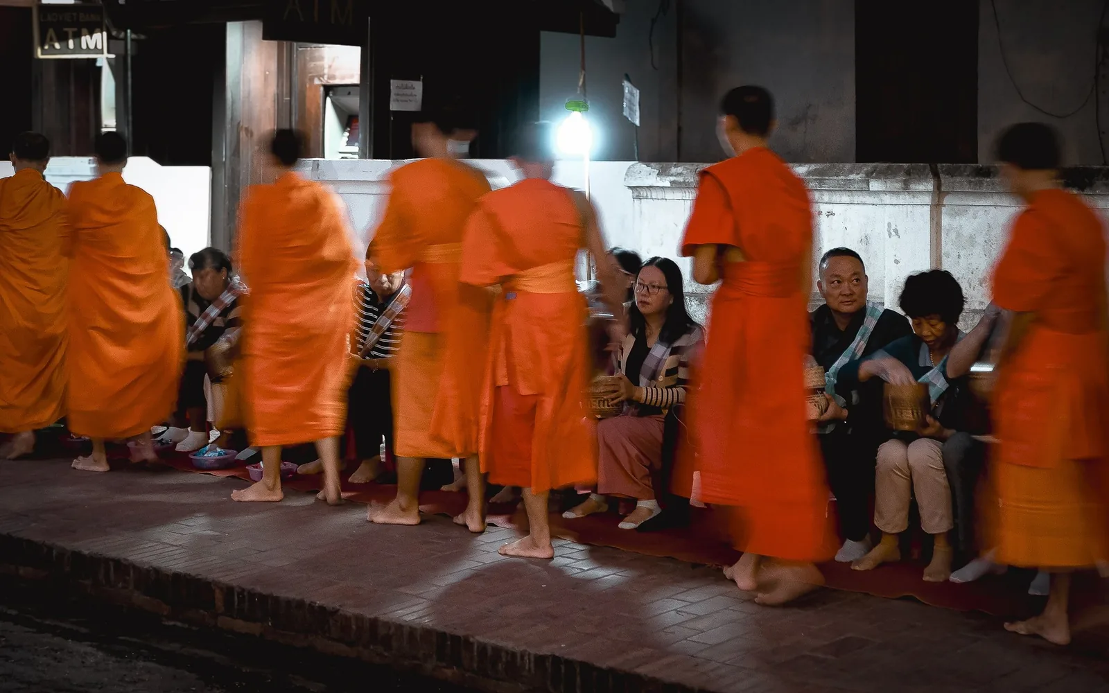 Barefoot monks in orange robes walk in a line at dawn as kneeling locals offer alms in Luang Prabang.