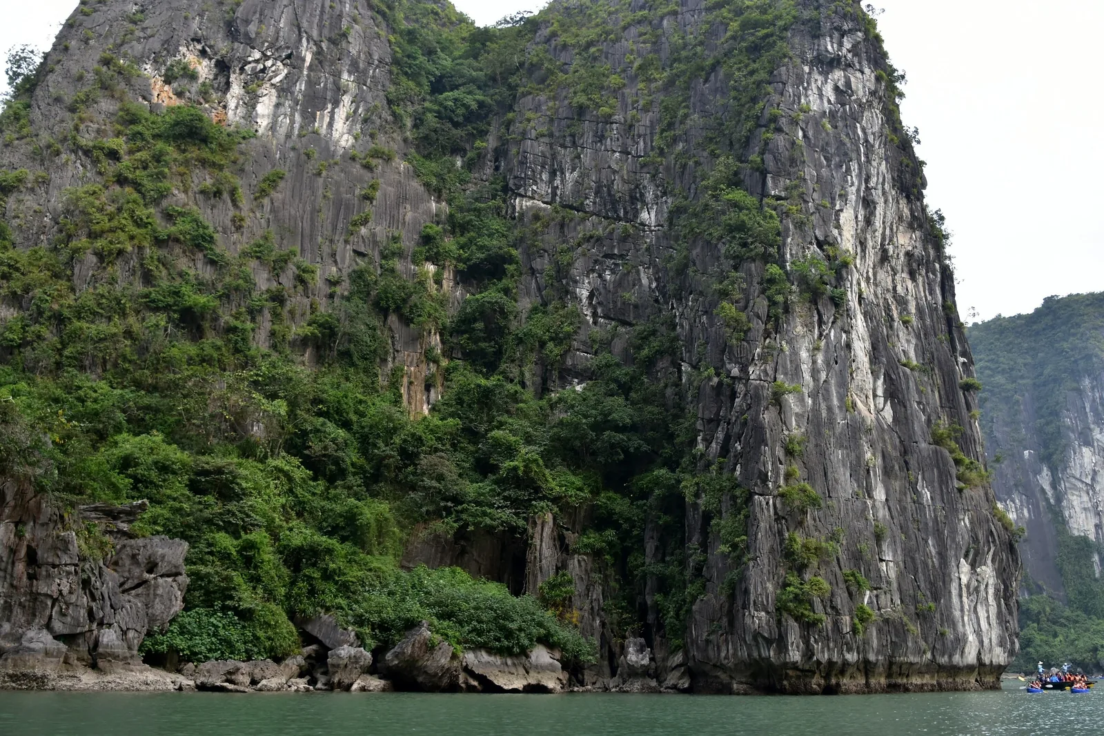 Towering limestone karst cliffs covered in greenery rise above the water in Ha Long Bay, with kayakers visible in the.