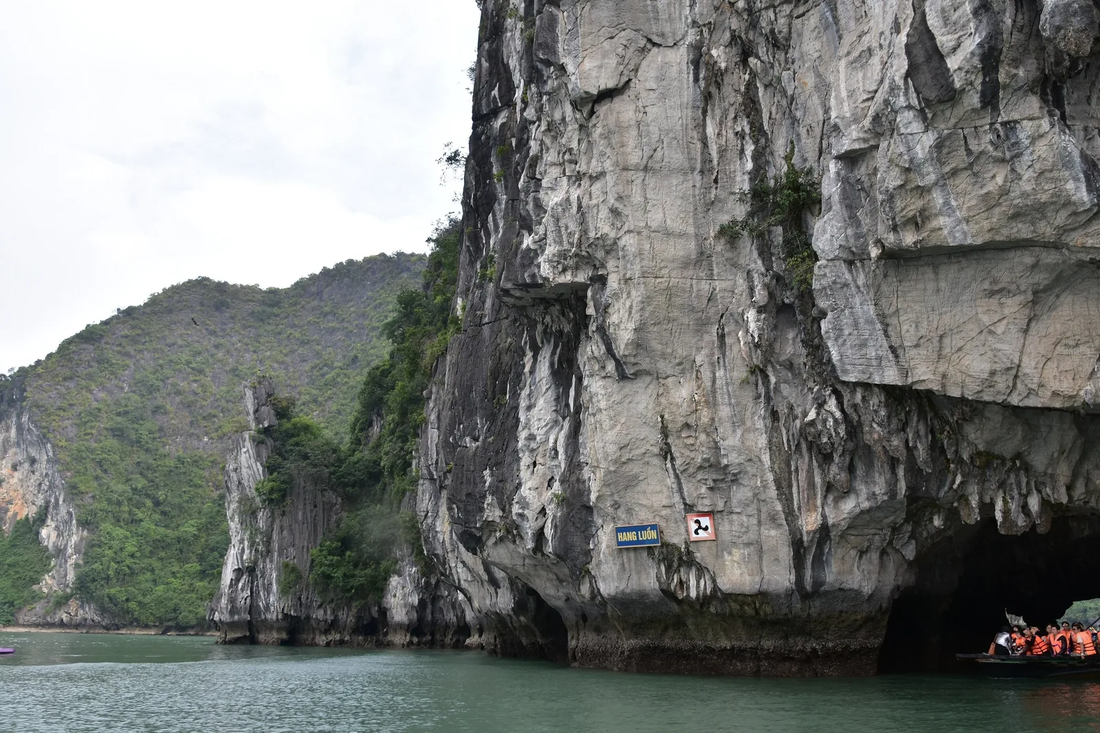 A limestone karst tunnel with a sign reading Hang Luon, small boats carrying tourists in life vests entering the cave in Ha.