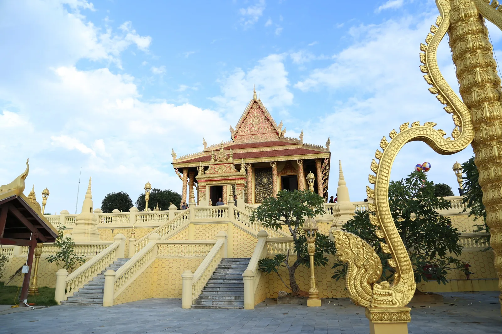 A golden Khmer-style temple pavilion with ornate naga sculptures and staircases stands under a blue sky in Vietnam.