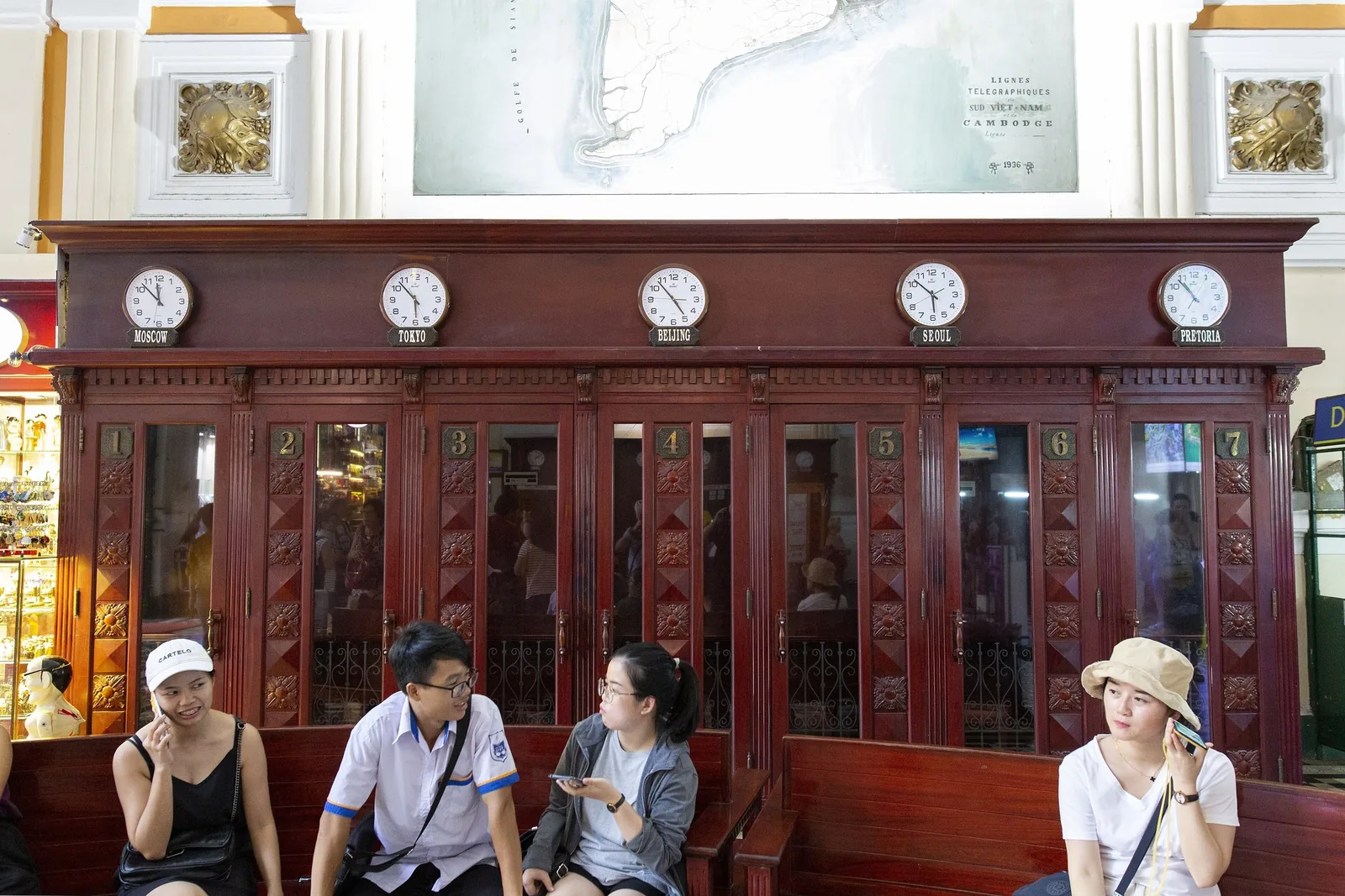 Visitors sit on a wooden bench below vintage world clocks and an old map inside Saigon Central Post Office in Ho Chi Minh.