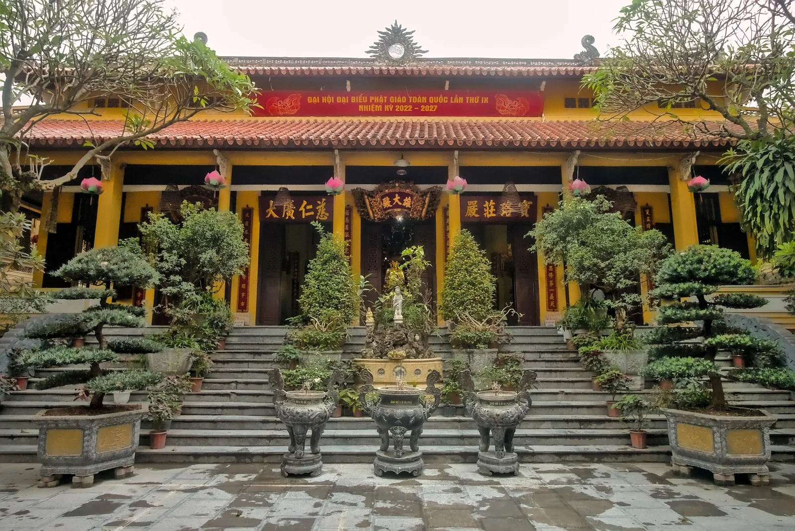 Yellow facade of Quan Su Temple in Hanoi with red-tiled roof, bonsai trees and stone steps leading to the entrance.