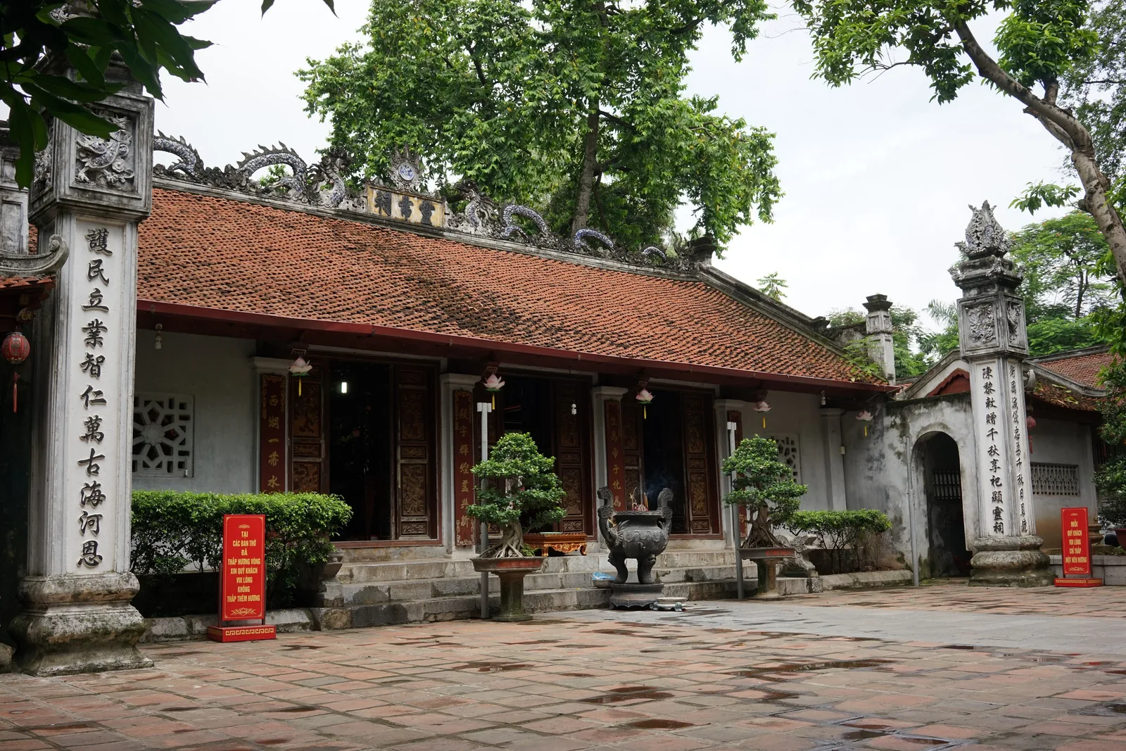 A traditional temple with red tiled roof and carved stone pillars stands in a courtyard in Hanoi.