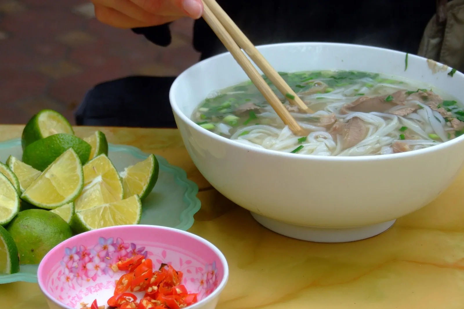 A steaming bowl of pho bo with beef and herbs, served with lime wedges and chopped chili on the side in Hanoi.