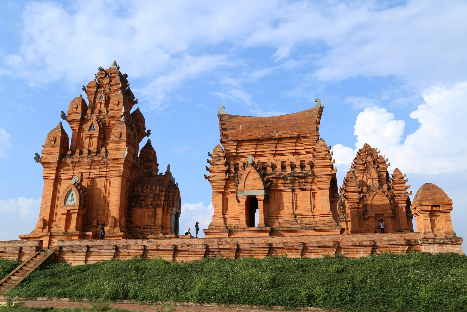 Ancient red brick Cham towers with intricate carvings stand on a grassy hill under a bright blue sky.