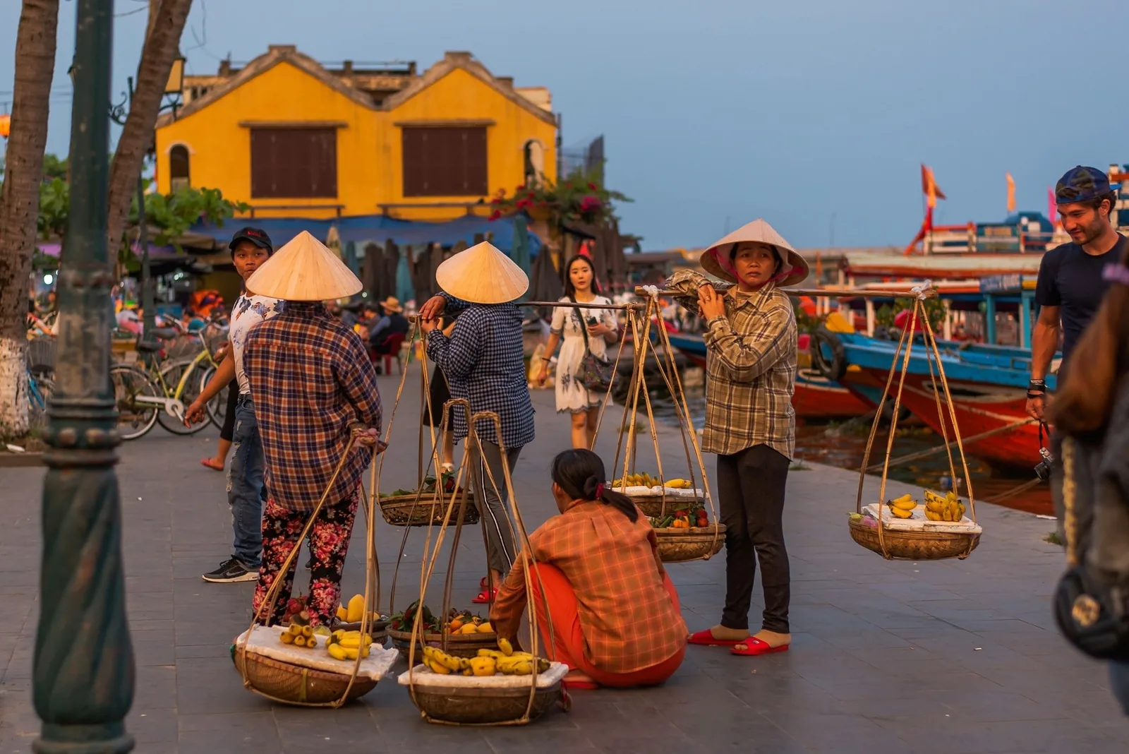 Fruit vendors with conical hats and shoulder baskets gather on the riverside promenade in Hoi An at dusk.