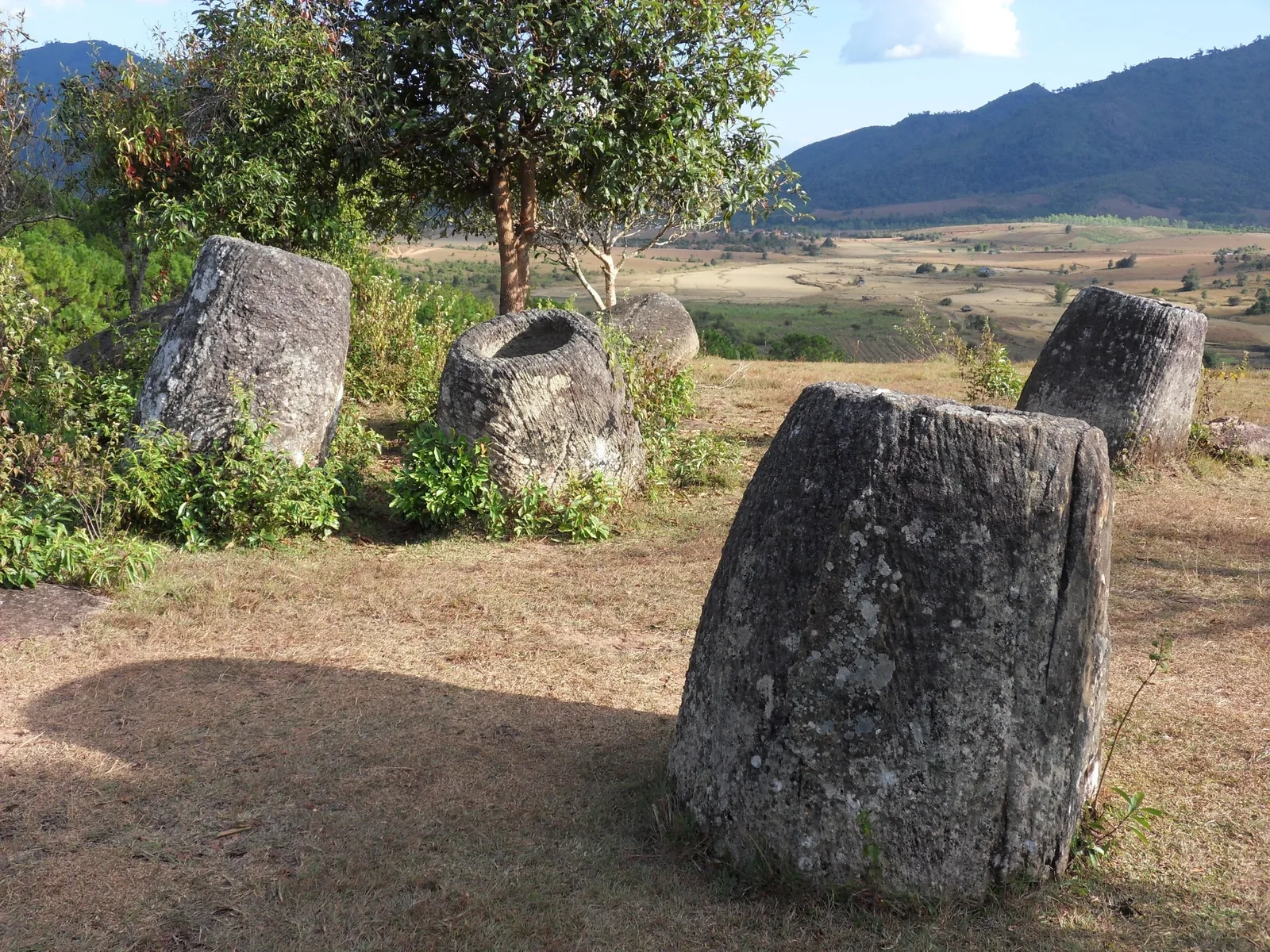 Ancient stone jars scattered on a grassy hillside overlooking valleys at the Plain of Jars, Laos.