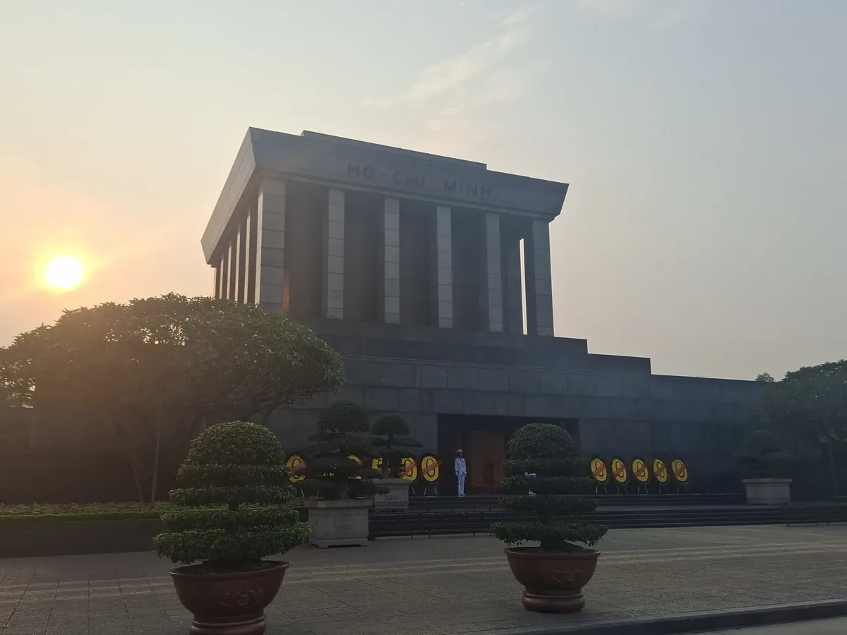 The Ho Chi Minh Mausoleum stands solemn in Hanoi at sunset, flanked by trimmed trees and a guard at the entrance.