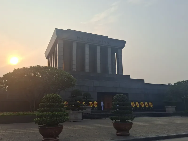 The Ho Chi Minh Mausoleum stands solemn in Hanoi at sunset, flanked by trimmed trees and a guard at the entrance.