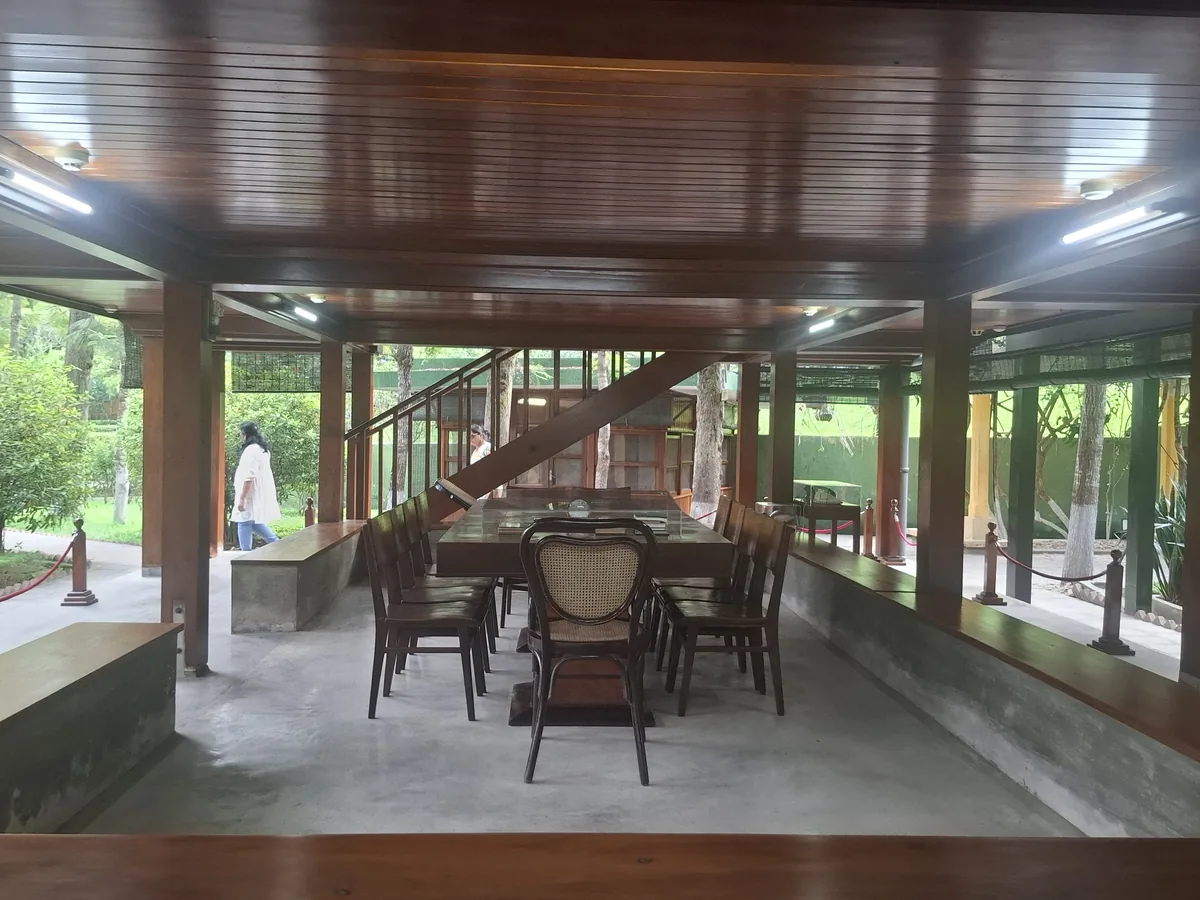 A wooden pavilion with a large table and chairs sits open-air within a garden setting in Hanoi.