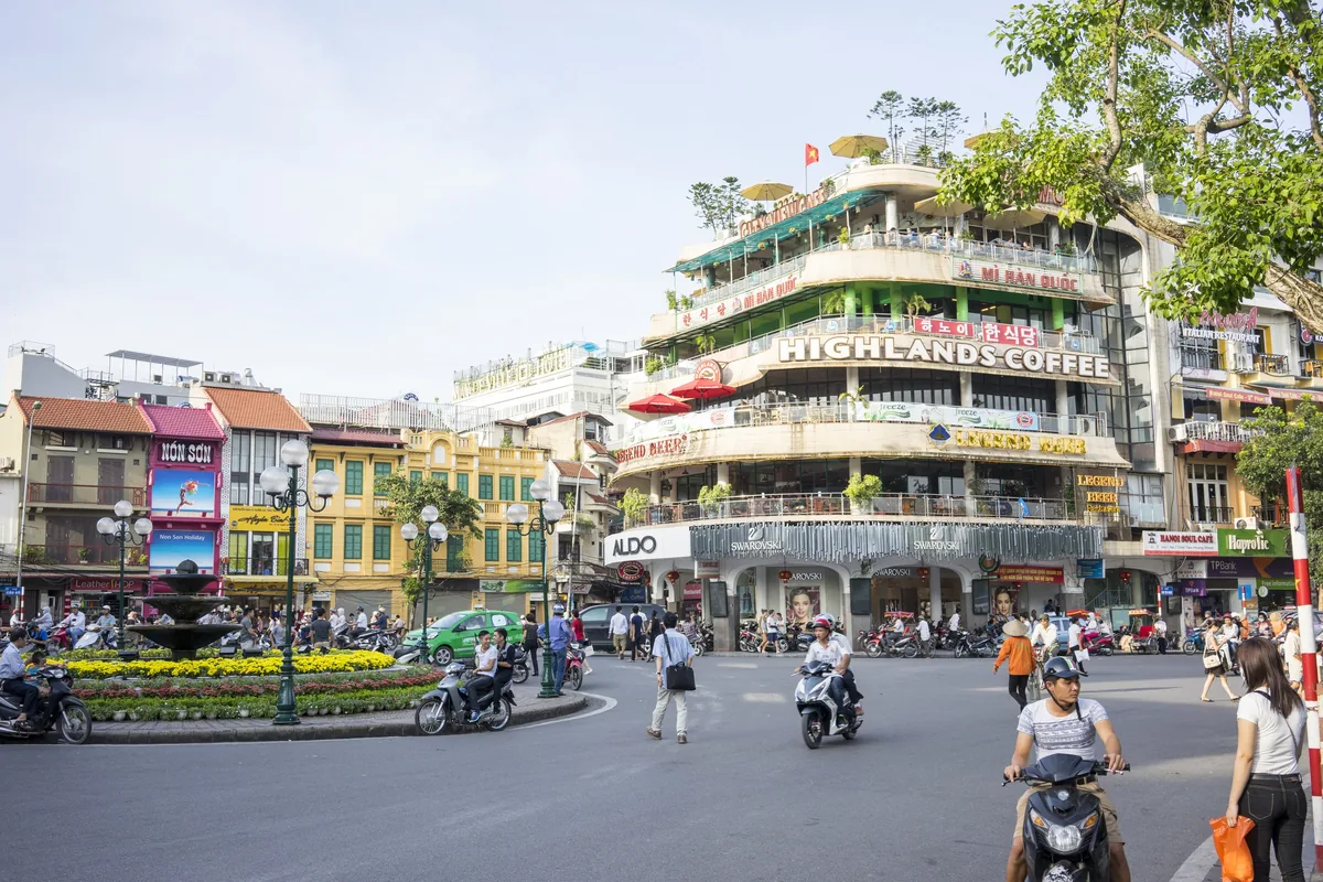 Motorbikes and pedestrians cross a busy roundabout beneath a curved corner building with the Highlands Coffee sign in Hanoi.