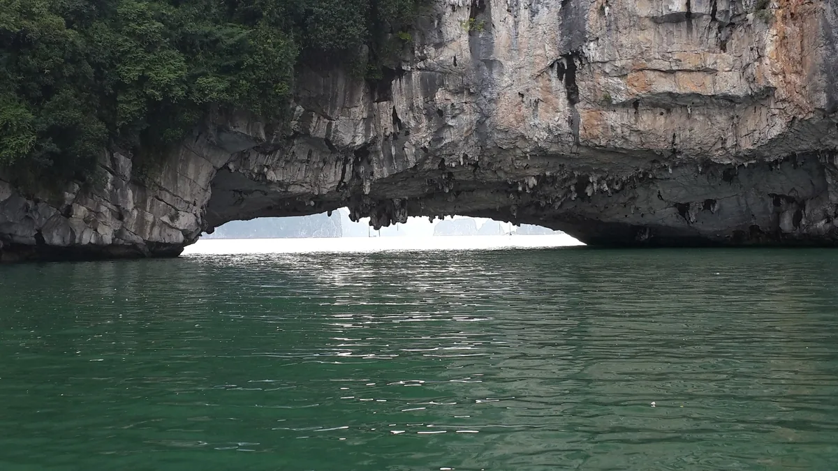 A natural stone arch carved into a limestone cliff rises above the emerald waters of Ha Long Bay.