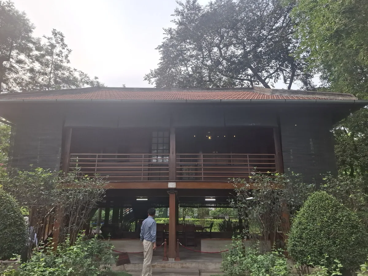 A visitor stands before Ho Chi Minh's stilt house, a wooden elevated structure surrounded by trees in Hanoi.