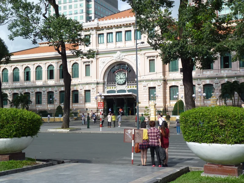 Colonial-era post office building with clock face and arched entrance in Saigon, framed by trees and modern skyscrapers.