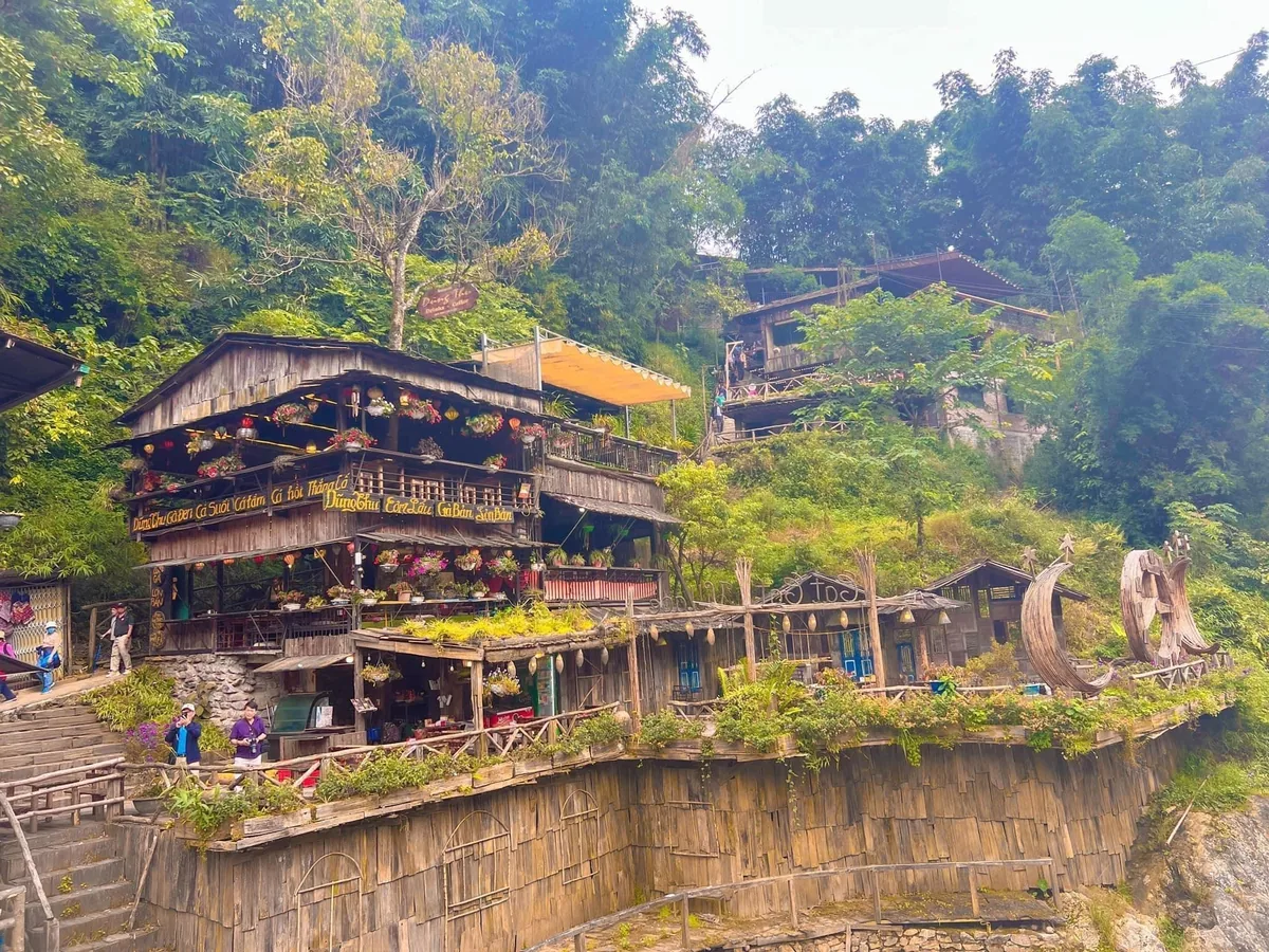 A wooden multi-level restaurant with flower decorations sits on a hillside in Cat Cat Village, surrounded by lush green.