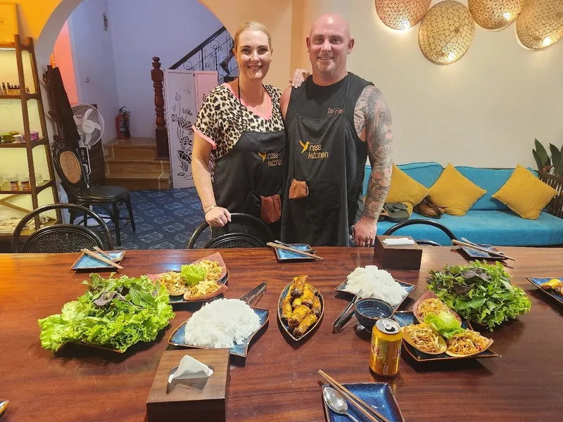 A smiling couple in aprons stand beside a table of Vietnamese dishes after a Hanoi cooking class.