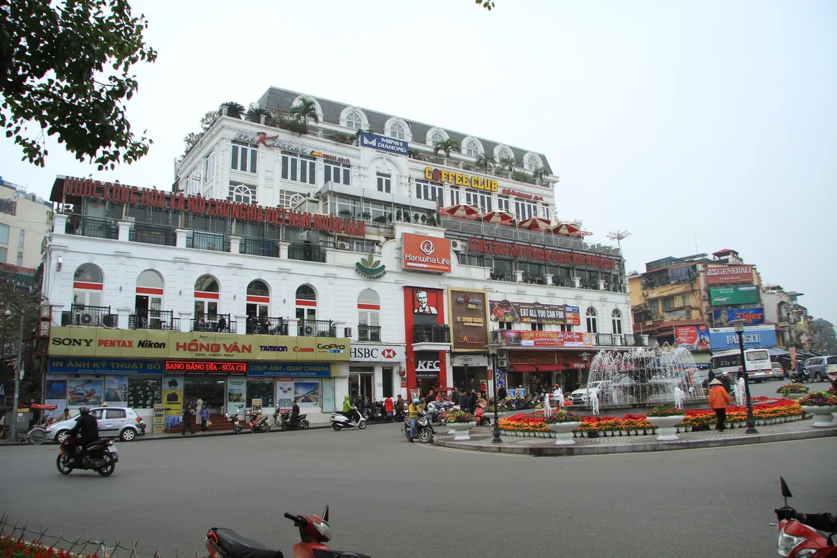 A busy Hanoi street corner with a colonial-style building covered in shop signs, a fountain, and motorbike traffic.
