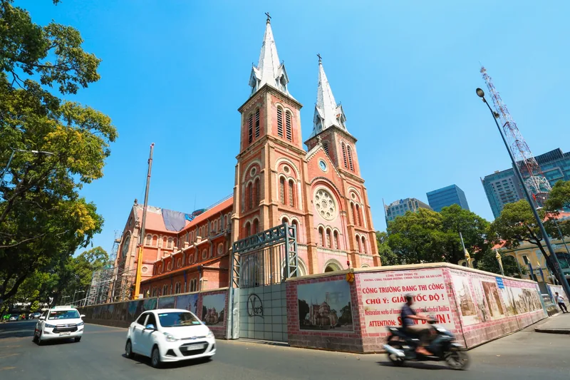 Twin spires of Notre Dame Cathedral rise above a busy street in Saigon, surrounded by construction hoarding and traffic.