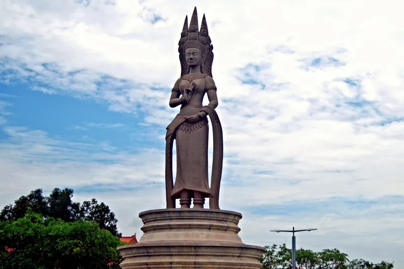 A tall Apsara statue with an ornate crown stands on a roundabout pedestal in Siem Reap under a partly cloudy sky.