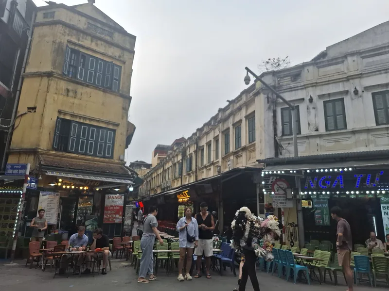 Colonial-style buildings line a busy street corner with outdoor cafes and pedestrians in Hanoi's Old Quarter.
