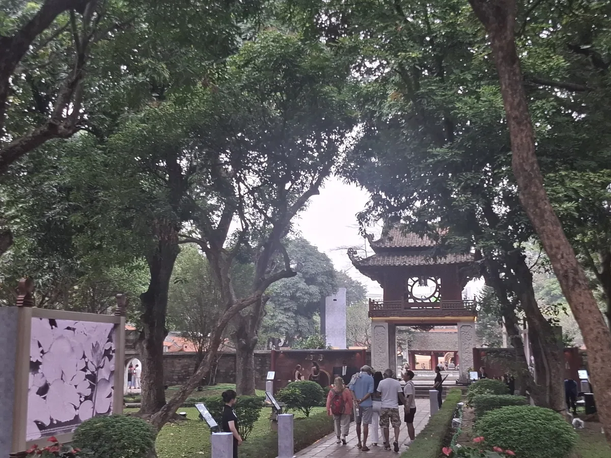 Visitors walk a shaded garden path toward a traditional wooden pavilion gate at a Hanoi temple complex.