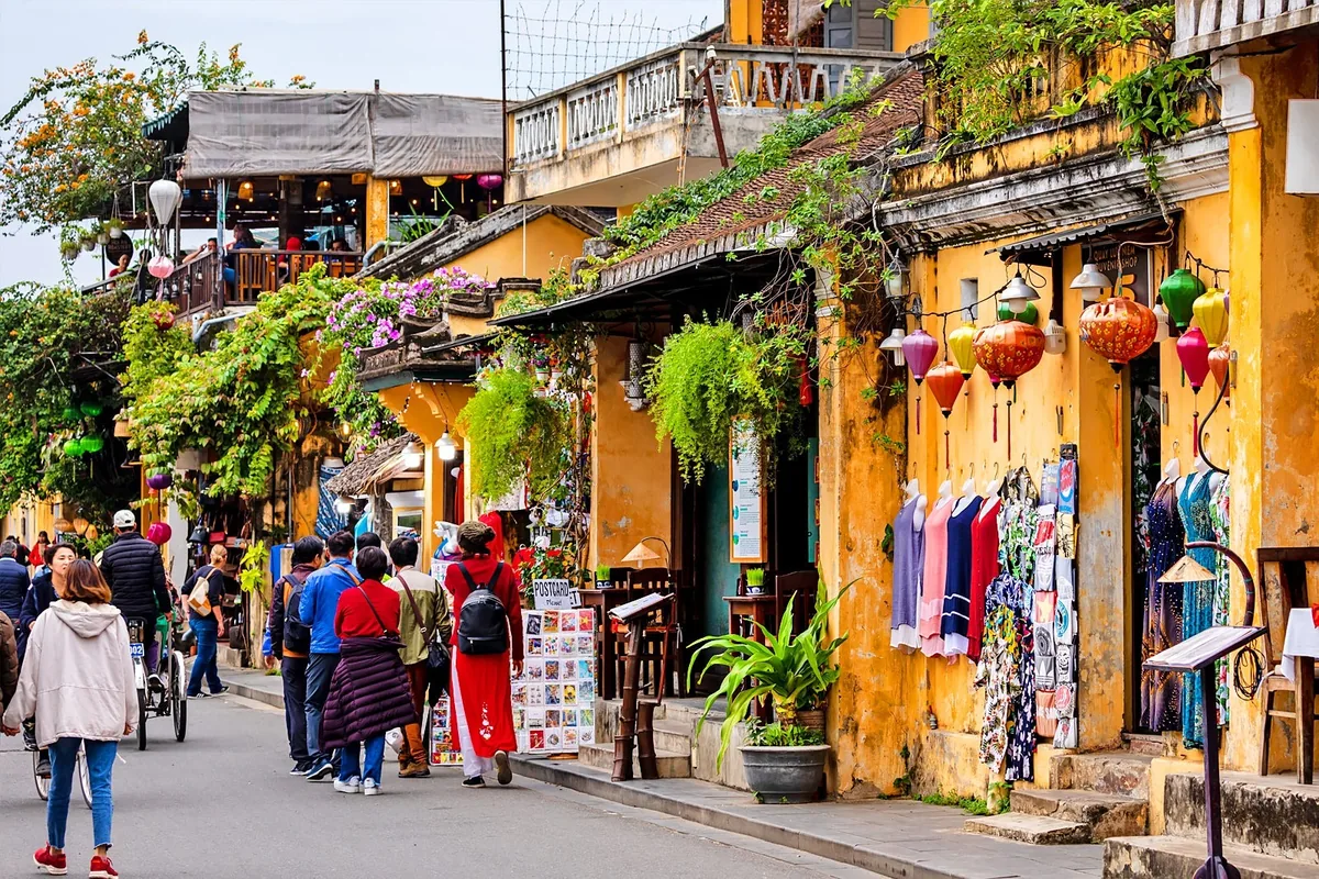 Colorful lanterns and yellow facades line a bustling street in Hoi An's old town as tourists stroll past shops.