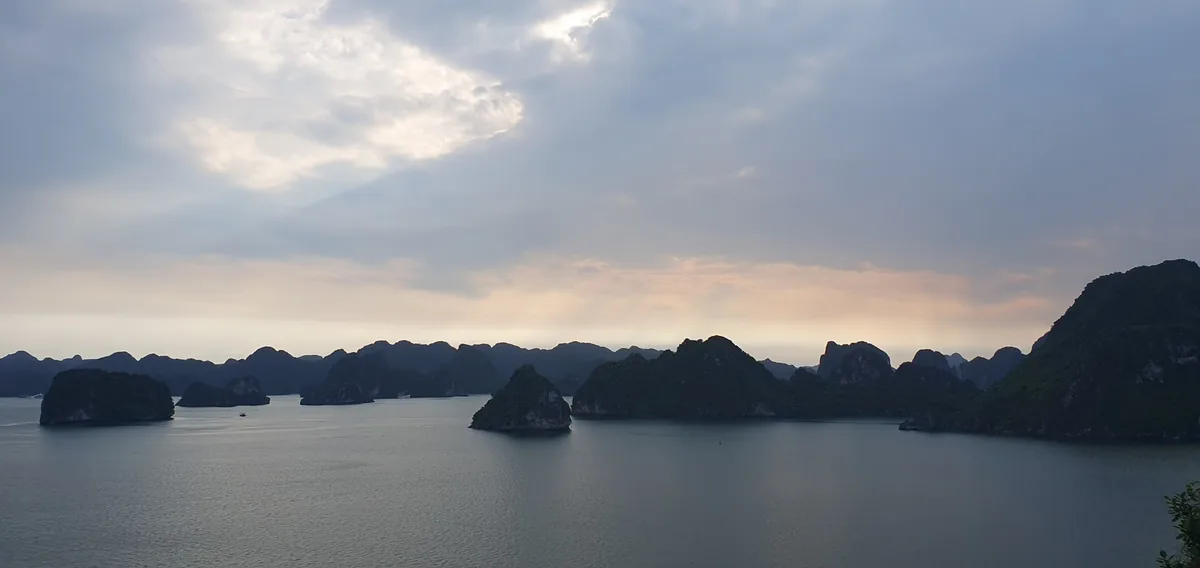 Limestone karsts rise from calm waters under a soft glowing sky at dusk in Halong Bay.