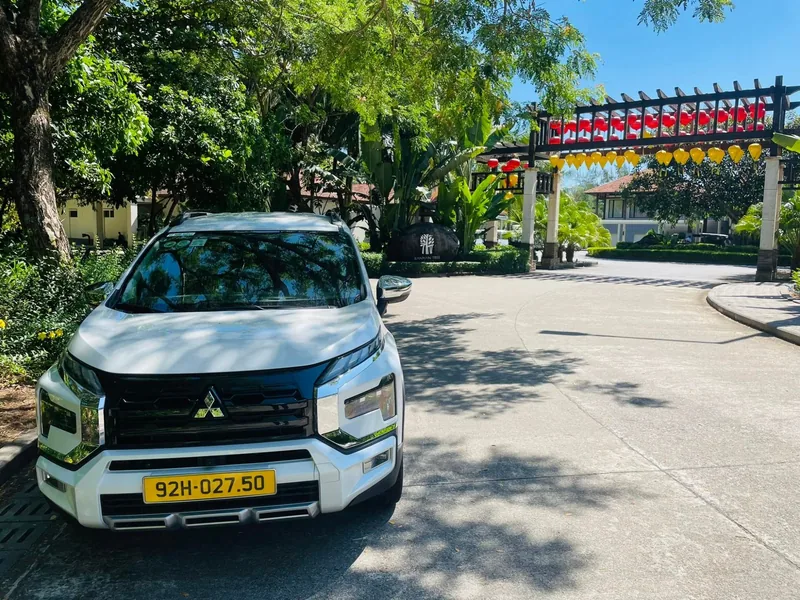 A white SUV with a yellow license plate parked on a shaded driveway leading to a resort entrance decorated with lanterns.