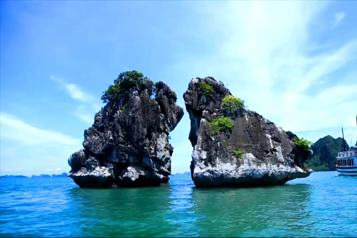 Two limestone karst rocks known as Trong Mai (Cock and Hen) rise from turquoise waters in Halong Bay with a cruise boat.