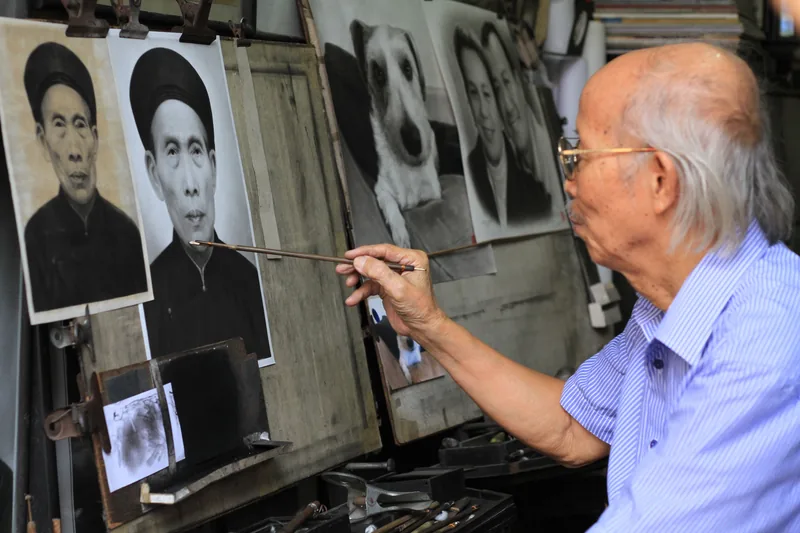 An elderly artist in Hanoi paints a portrait, surrounded by charcoal drawings hanging in his studio.