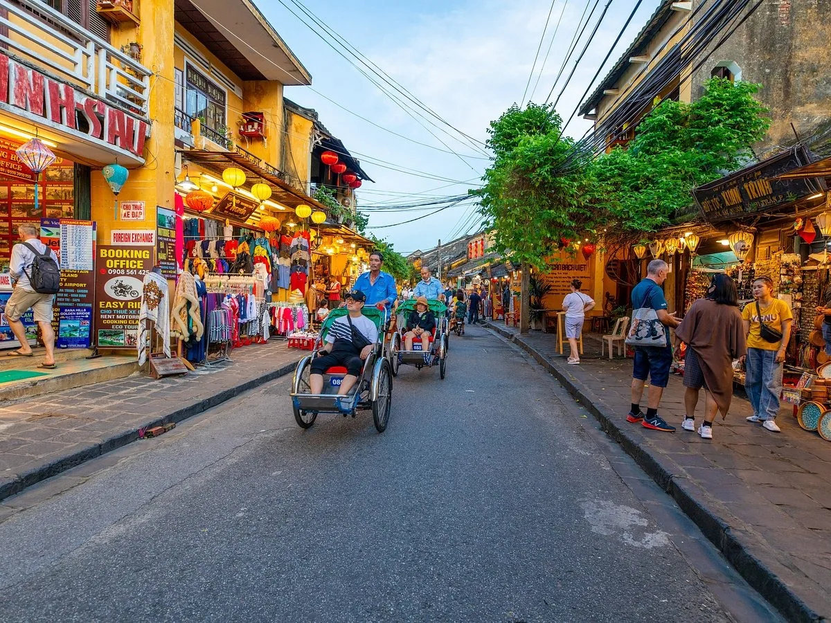 Cyclos carry tourists along a lantern-lined street in Hoi An's ancient town at dusk, flanked by shophouses and market stalls.