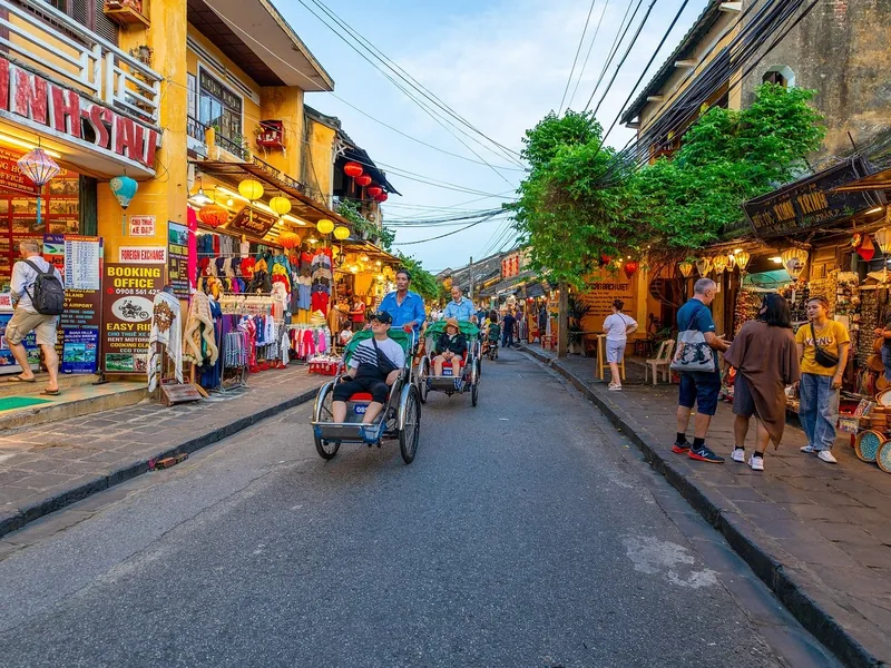 Cyclos carry tourists along a lantern-lined street in Hoi An's ancient town at dusk, flanked by shophouses and market stalls.