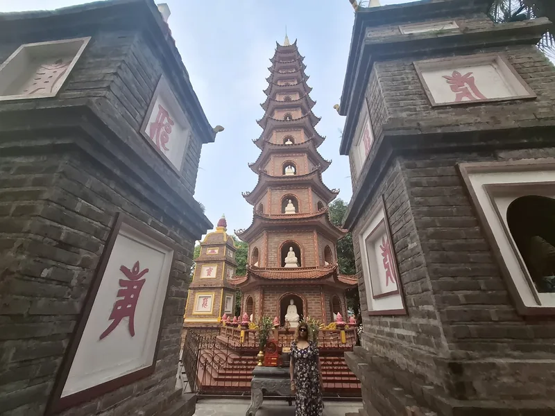 A woman stands before the multi-tiered brick tower of Tran Quoc Pagoda in Hanoi, flanked by ancient stone walls.