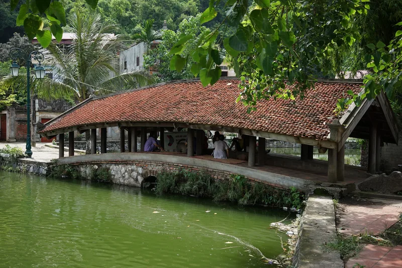 A traditional covered wooden bridge with a red tiled roof arches over a green pond near Hanoi.