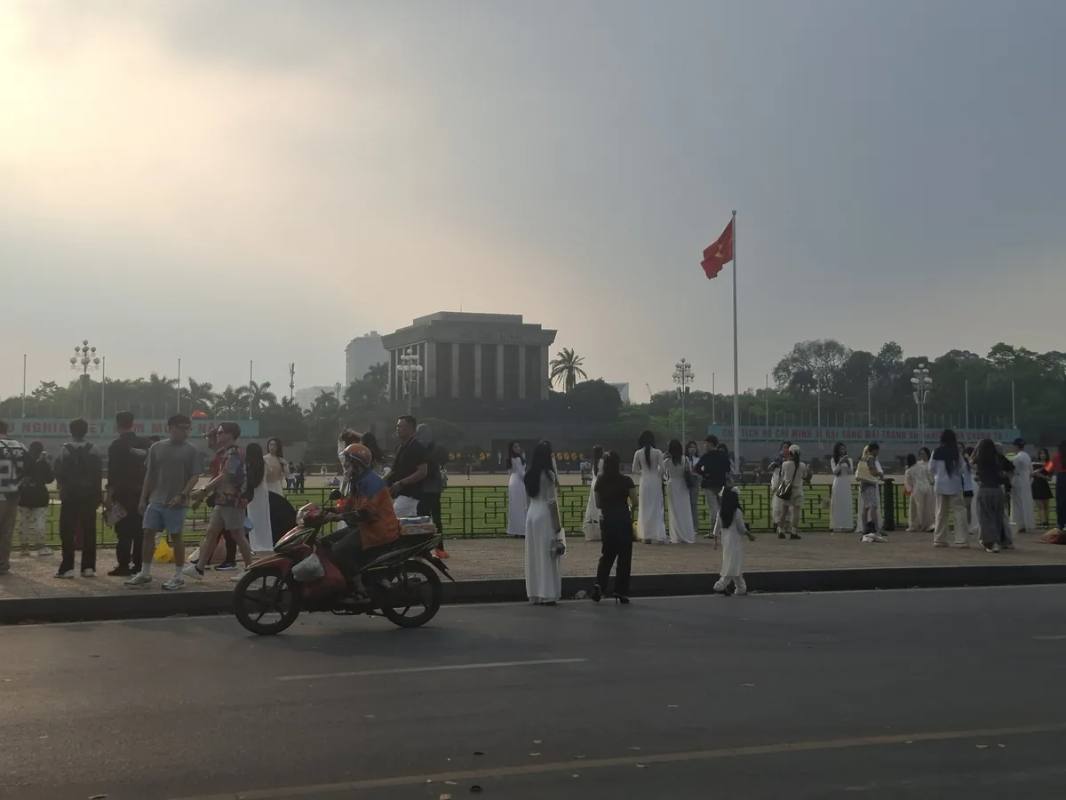 People gather near the mausoleum building in Ba Dinh Square, Hanoi, with a red flag flying and women in white ao dai in the.