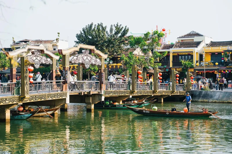 A lantern-decorated bridge crosses the river beside colorful shophouses in Hoi An's old town, with boats floating below.