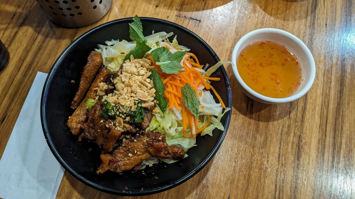 A bowl of Vietnamese vermicelli noodles topped with grilled meat, herbs, carrots and peanuts, served with dipping sauce.