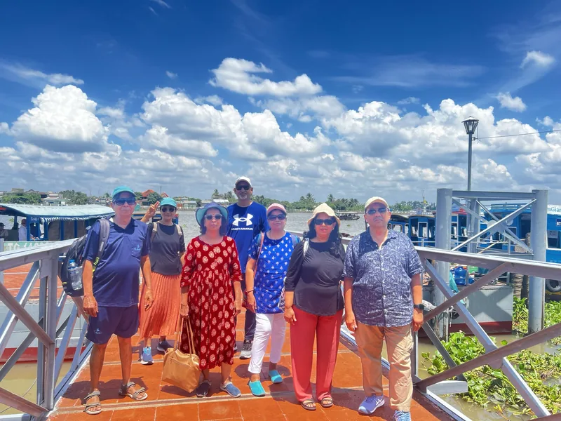 A group of tourists poses on a wooden pier along a wide river in the Mekong Delta under a bright blue sky.