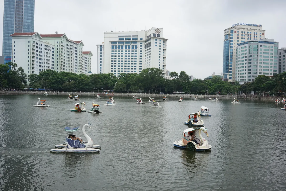 Swan-shaped pedal boats float on a lake in Hanoi with high-rise buildings lining the shore behind trees.