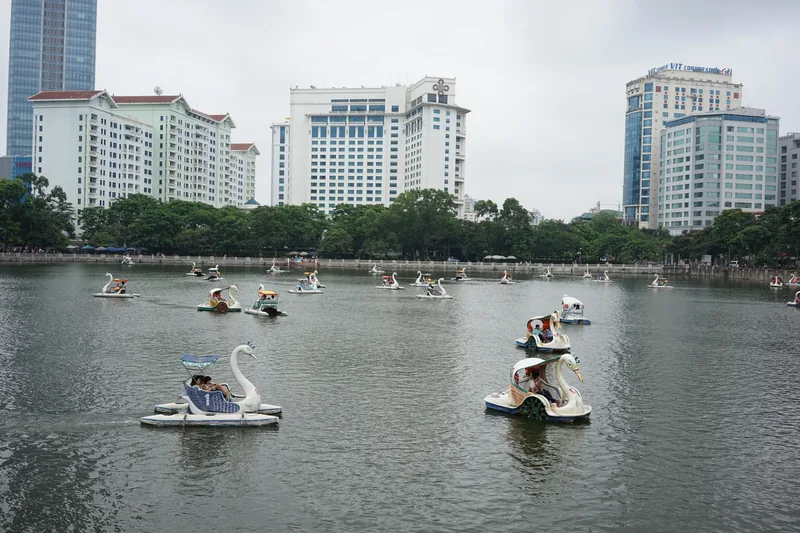 Swan-shaped pedal boats float on a lake in Hanoi with high-rise buildings lining the shore behind trees.