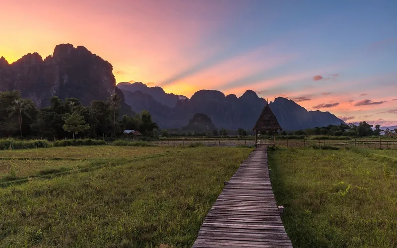 A wooden walkway leads across green rice fields to a thatched hut beneath karst mountains at sunset in Vang Vieng.
