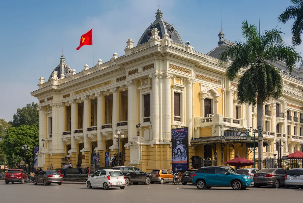 The colonial-era Hanoi Opera House stands under a clear blue sky, flying the Vietnamese flag with cars parked in front.