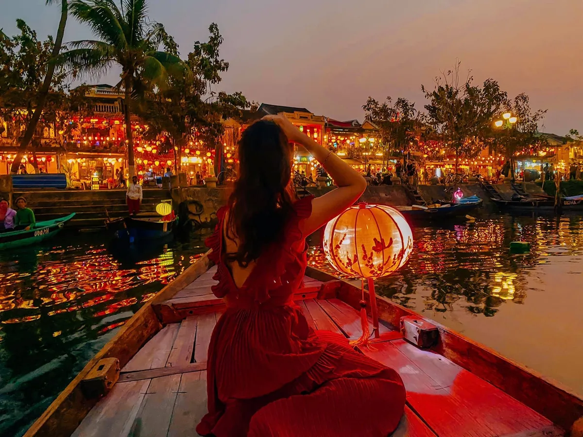 A woman in a red dress sits on a boat lit by a lantern, gazing at Hoi An's glowing riverside old town at dusk.